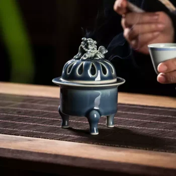 Blue ceramic incense burner with steam rising, on a wooden mat, while a hand holds a cup in the background.